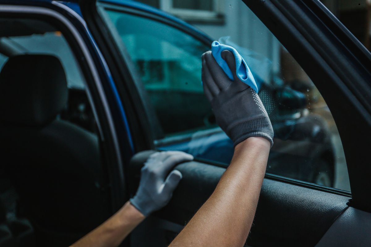 A person wearing gray gloves cleans the inside of a car window with a blue cloth, focused on removing dirt and smudges.