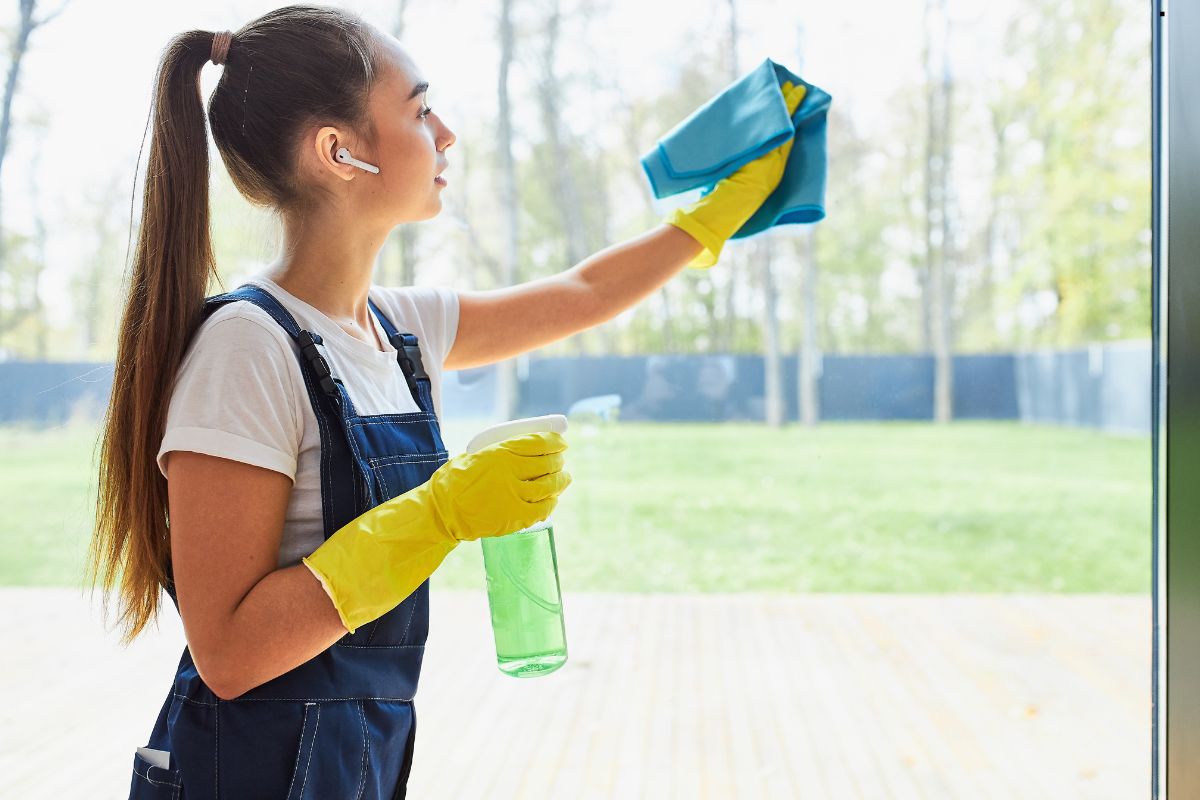 A person wearing yellow gloves wipes down a window with a blue cloth, holding a green spray bottle, surrounded by greenery.