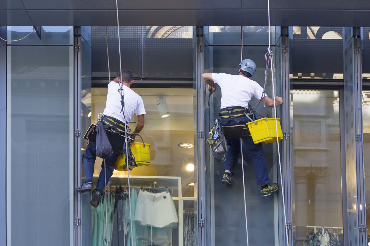 Two men using squeegees to clean windows on a tall building, showcasing teamwork and attention to detail. 